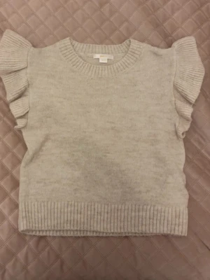 Beige stickad topp med volangärm - Supersöt beige stickad topp med korta volangärmar och rund halsringning. Ribbstickad kant nedtill och vid ärmslut ger en snygg finish. Perfekt för dig som vill ha något gulligt och trendigt i garderoben.
