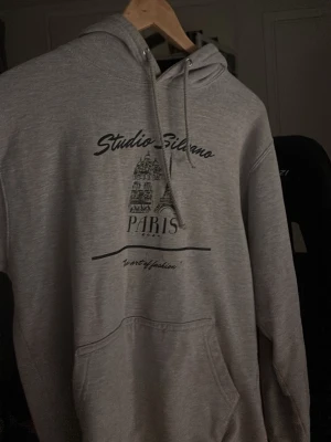 Grå hoodie Studio Silvano Paris - Grå hoodie med tryck från Studio Silvano. Framsidan har svart text och motiv av Paris med Eiffeltornet. Klassisk huva med dragsko och stor magficka. Perfekt för en chill och avslappnad stil.