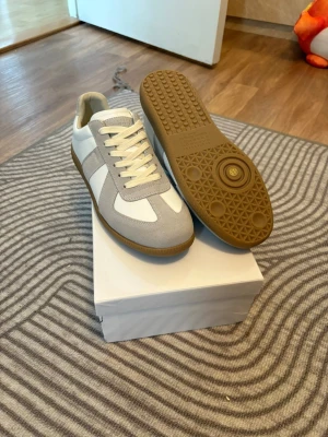 Vita maison margiellas sneakers - Stilrena sneakers i vitt och ljusgrått med klassisk retrodesign. Ovandelen är i en mix av slätt läder och mocka, och sulan är i gummi med mönstrad undersida för bra grepp. Perfekta för dig som gillar minimalistisk och tidlös stil.
