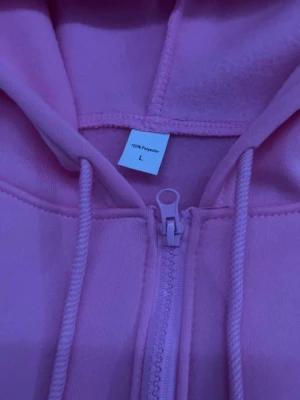 Rosa zip hoodie med logga - Säljer en rosa hoodie med dragkedja och huva. Tröjan har snören i huvan och en broderad svart logga på bröstet. Tillverkad i 100% polyester och har en mjuk insida. Perfekt för dig som vill ha en chill och snygg look.