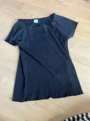Svart ribbad topp från Lindex - Säljer en svart ribbad topp från Lindex med korta ärmar och stretchigt material. Toppen har en lätt vågig kant vid halsringning, ärmslut och nederkant. Perfekt till dig som gillar en enkel men snygg stil. 