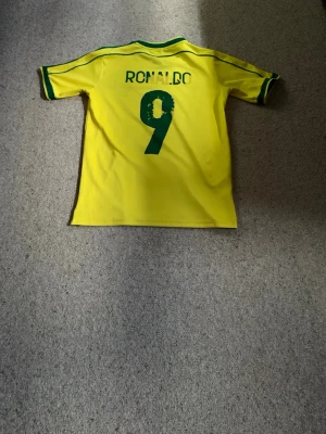 Brasilien Ronaldo 9 matchtröja Nike - Gul Brasilien matchtröja från Nike med gröna detaljer, nummer 9 och namnet Ronaldo på ryggen. Tröjan har officiellt landslagsemblem, gröna ränder på ärmarna och texten 'FIFA World Cup France 98' på bröstet. Perfekt för fotbollsfans! Passar 150 cm