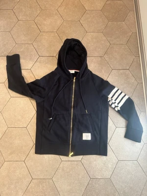 Marinblå Thome Browne hoddie  - Snygg marinblå Thome Browne hoodie med dragkedja och huva. Ena ärmen har fyra vita ränder och en patch på magen. Insidan har en randig ficka i rött, blått och vitt. Perfekt för en avslappnad streetwear-look. 