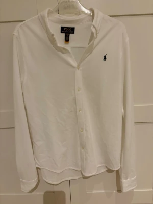 Vit skjorta från Polo Ralph Lauren - Klassisk vit piké skjorta från Polo Ralph Lauren med knappar framtill och loggan på bröstet. Skjortan har lång ärm. Använd 1 gång och köpt på Kidsbrandstore. XL i juniorstorlek!