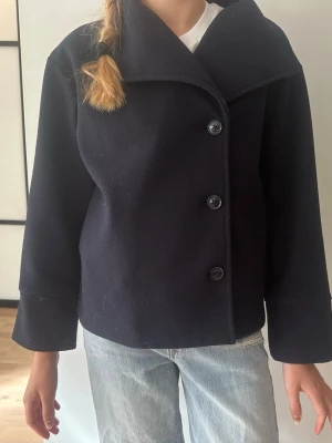 Mörkblå ullrock med stora knappar - Stilren mörkblå rock i ullmix med tre stora svarta knappar framtill och bred krage. Rak passform och långa ärmar ger en klassisk look. Perfekt för dig som vill ha en enkel men snygg jacka till hösten eller vintern.