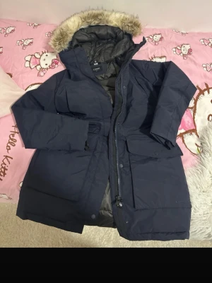 Mörkblå parkas från Everest - Snygg mörkblå parkas från Everest med avtagbar fuskpäls på huvan och quiltat foder. Jackan har dragkedja, stora fickor framtill och justerbar midja. Perfekt för kalla dagar och riktigt mysig att bära.