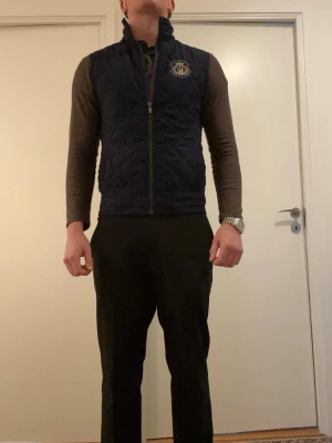 Mörkblå quiltad dunväst - Snygg mörkblå dunväst med quiltat mönster och hög krage. Västen har dragkedja framtill och en broderad logga på vänster bröst. Perfekt att slänga över en hoodie eller tröja för extra värme och stil. (Är 186 lång)