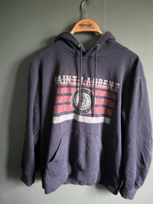 Mörkblå hoodie från Saint Laurent - Mörkblå hoodie från Saint Laurent med tryck i vitt och rött framtill samt texten 'SAINT LAURENT'. Klassisk känguruficka och justerbar huva med snören. Mjuk insida och avslappnad passform, perfekt för en cool och bekväm stil. Den är i storlek M men sitter mer som S.