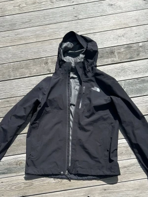 Svart regnjacka från The North Face - Snygg svart regnjacka från The North Face med huva och dragkedja framtill. Jackan har en bröstficka med dragkedja och justerbar huva med snörning. Perfekt för regniga dagar och outdoor-äventyr.