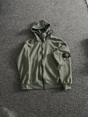 Grön hoodie med dragkedja Ston Island - Snygg grön hoodie från Ston Island med dragkedja framtill och klassisk patch på vänster ärm. Huva med dragsko och fickor på sidorna. Perfekt för en chill och stilren look.