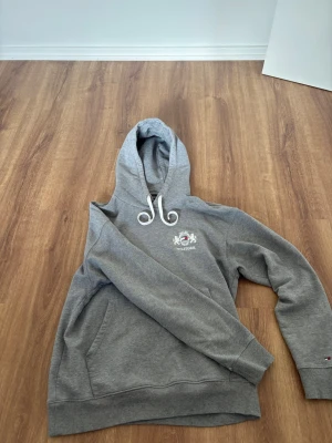 Grå hoodie från Tommy Hilfiger - Snygg grå hoodie från Tommy Hilfiger med broderad logga och text på bröstet. Klassisk huva med vita dragsnören och känguruficka framtill. Tillverkad i mjukt bomullsmaterial och har ribbade muddar vid ärmslut och nederkant. Pris diskuterbart vid snabb affär.