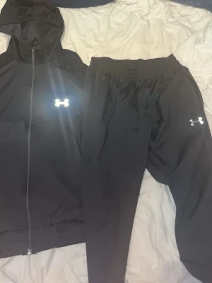 Under armour  - Snyggt svart träningsset från Under Armour med matchande hoodie och byxor. Hoodien har dragkedja och logga på bröstet, byxorna har resår i midjan och logga på benet. Perfekt för gymmet eller chill dagar.