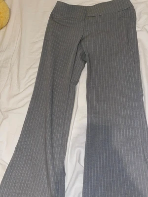 Grå kostymbyxor med smala ränder - Snygga grå kostymbyxor med vita smala kritstrecksränder och utsvängda ben. Byxorna har en klassisk midja och är sydda i ett mjukt tyg som ger en dressad look. Perfekta för dig som gillar stilrena och trendiga plagg.