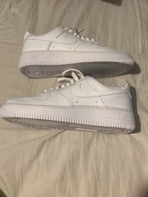 Nike Air Force 1 vita sneakers - Klassiska helvita Nike Air Force 1 sneakers i läder med lågt skaft och ikonisk sula. Swoosh-logga på sidorna, AIR-detalj på sulan och perforerad tå för extra ventilation. Perfekta för dig som vill ha en clean och tidlös look. Utan orginalkartong
