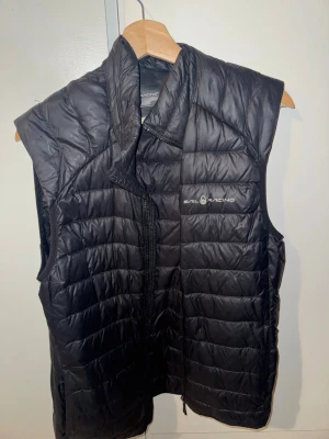Svart dunväst från Sail Racing - Snygg svart dunväst från Sail Racing med quiltad design och hög krage. Västen har dragkedja framtill och logga på bröstet samt en diskret logga i nacken. Perfekt för lager-på-lager och håller dig varm under kyliga dagar.