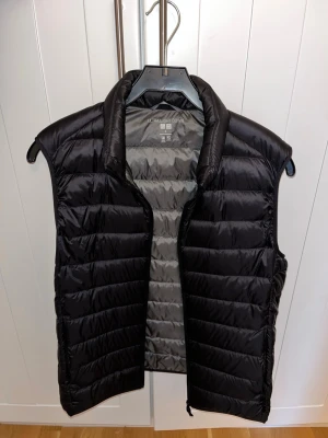 Svart Väst Uniqlo - Snygg svart väst. Västen är lätt och smidig, Insidan är grå och materialet är glansigt, vilket ger en modern look. Passar till många olika stilar och är enkel att matcha.