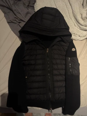 Svart Moncler Cardigan - Svart Moncler Cardigan med stickade ärmar och stor huva. Jackan har dragkedja framtill och en ficka med dragkedja på ena ärmen. Perfekt för kalla vinterdagar och har en modern, sportig look.