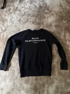 Svart Peak Performance tröja  - Klassisk svart sweatshirt från Peak Performance med vit text på bröstet. Tröjan har rund halsringning, långa ärmar och ribbade muddar vid ärmslut och nederkant. Perfekt för en avslappnad och stilren look.