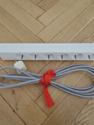 Vit grenuttag med cirka 3,5 msladd - Praktiskt vitt grenuttag med sex uttag och extra lång grå sladd. Perfekt för att koppla in flera elektronikprylar på samma ställe. Sladden är hopbunden med ett rött band för enkel förvaring. Har 6 jordade uttag.