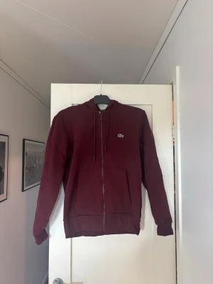 Vinröd Zip från Lacoste - Snygg vinröd ziphoodie från Lacoste med dragkedja framtill och klassisk krokodil-logga på bröstet. Hoodien har långa ärmar, fickor fram och justerbar huva med snören. Perfekt för en chill och stilren look.