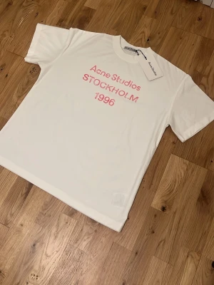 Vit Acne Studios t-shirt med tryck - Säljer en vit t-shirt från Acne Studios med rosa texttryck på bröstet där det står 'Acne Studios STOCKHOLM 1996'. Klassisk passform och rund halsringning. Perfekt för dig som gillar stilrena och trendiga plagg.