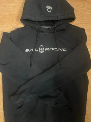Svart hoodie från Sail Racing - Snygg svart hoodie från Sail Racing med logga tryckt på bröstet och en mindre logga på huvan. Klassisk känguruficka framtill och justerbar dragsko i huvan. Perfekt för en avslappnad och sportig stil.