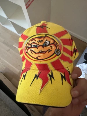 Gul keps med tecknad sol och blixtar - Snygg gul keps med röd och svart grafik, stor tecknad sol med ansikte framtill och blixtar på skärmen. Baksidan har svart panel med tryckt hjälmfigur och justerbart spänne med texten 'www.daring.com'. Perfekt för dig som gillar unika accessoarer.