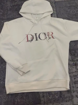 Vit hoodie med blommig tryck - Snygg vit hoodie med stor huva och dragsko. På bröstet med blommigt mönster i rosa, lila och svart. Hoodien har ribbade muddar och är gjord i mjukt material, perfekt för en chill och trendig look.