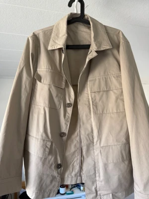 Beige overshirt med fickor - Stilren beige overshirt med fyra stora fickor framtill och knäppning med bruna knappar. Klassisk krage och raka ärmar. Tillverkad i ett lätt bomullsliknande material som passar perfekt för lager på lager. Enkel och clean design utan synligt märke.