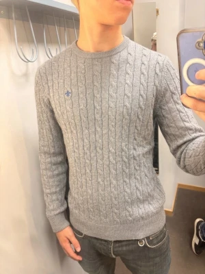 Grå kabelstickad tröja Morris - Snygg grå kabelstickad tröja från Morris i 100% bomull. Tröjan har rund halsringning, ribbade muddar och en liten blå logga broderad på bröstet. Perfekt för dig som gillar klassisk och stilren design.