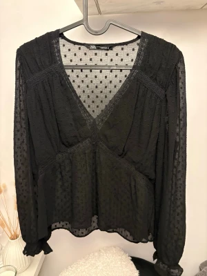 Svart transparent blus från Zara - Snygg svart blus från Zara i transparent mesh med diskret prickigt mönster. Djup V-ringning och långa ärmar med volangdetalj vid ärmslut. Perfekt för lager på lager och ger en trendig look till din outfit.