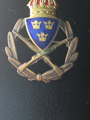 Svenska försvarsmakten brosch med blått emaljsköld stämplad "585" och "14k" - Unik militär brosch i metall med blå emaljsköld prydd med tre kronor, korsade gevär och lagerkrans. Ovanpå skölden sitter en guldfärgad krona med röda detaljer. Perfekt som accessoar eller samlarobjekt. Broschen är stämplad som att den vore i 14karat guld,  alltså "585" & "14k' men jag har guldtestat den och den gav inget utslag.