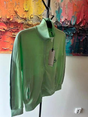 Palm angels  - Snygg mintgrön ziptröja med hög krage och svarta ränder längs ärmarna. Tröjan har dragkedja framtill, två fickor med dragkedja och ribbade muddar vid ärmslut och nederkant. Perfekt för en sportig och avslappnad stil.