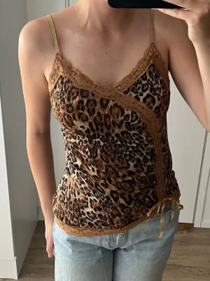 Glittrig leopard topp - Snygg leopardmönstrad linnetopp med tunna axelband och spetsdetaljer i brunt. Toppen har omlottkänsla framtill och en liten rosett i sidan.