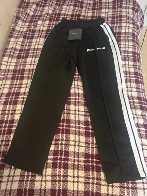 Palm Angels svarta mjukisbyxor - Palm Angels svarta mjukisbyxor med vita sidoränder och logga på vänster lår. Byxorna har resår i midjan och raka ben. Perfekta för en avslappnad och trendig streetwear-look.