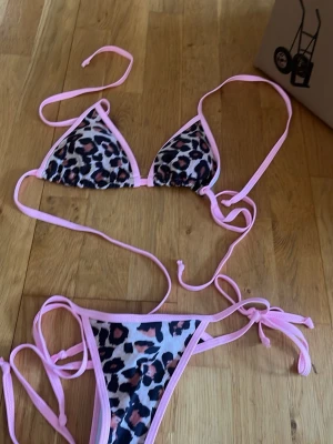 Leopardmönstrad bikini med rosa kant - Snygg trekantsbikini med leopardmönster och ljusrosa kanter. Både överdel och underdel har knytband för justerbar passform. Perfekt för sommaren och strandhäng!