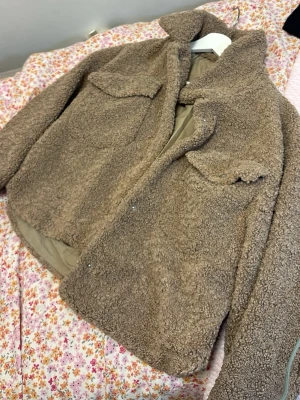Beige teddyjacka från Zara - Mysig beige teddyjacka från Zara med två stora bröstfickor och knappar framtill. Jackan har en hög krage och är tillverkad i fluffigt teddy-material som håller dig varm. Perfekt för dig som vill ha en trendig och bekväm look under kyliga dagar.