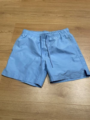 Ljusblå shorts med snörning - Säljer ett par ljusblå shorts med elastisk midja och snörning framtill. Shortsen har en enkel och clean design med sidofickor och är perfekta för varma dagar. Tillverkade i ett lätt och bekvämt material.