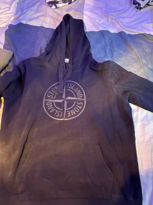 Svart hoodie från Stone Island - Säljer en svart hoodie från Stone Island med stor logga på bröstet och klassisk patch på ärmen. Hoodien har huva med dragsko och en stor magficka. Perfekt för dig som gillar streetwear och vill ha något stilrent och bekvämt.