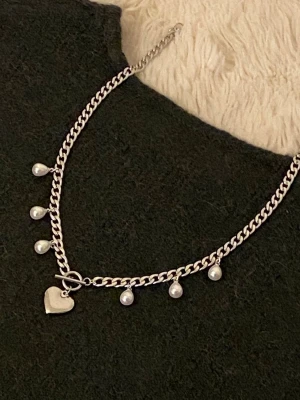 Silverfärgat halsband med pärlor och hjärta - Snyggt silverfärgat halsband med sex vita pärlhängen och ett hjärtformat hänge. Kedjan har en chunky design och stängs med ett dekorativt T-lås. Perfekt för att ge din outfit en trendig touch.