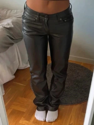 Svarta raka byxor i skin - Snygga svarta byxor i skin med rak passform och låg midja. Klassisk femficksmodell med dragkedja bak och bälteshällor. Perfekta för en trendig och edgy look.