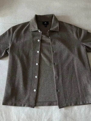 Mörk grön skjorta med kort ärm och knappar - Säljer en stilren mörk grön skjorta från H&M med korta ärmar och vita knappar framtill. Skjortan har klassisk krage och är gjord i ett mjukt, strukturerat material som känns skönt mot huden. Perfekt för dig som gillar enkel och clean stil.
