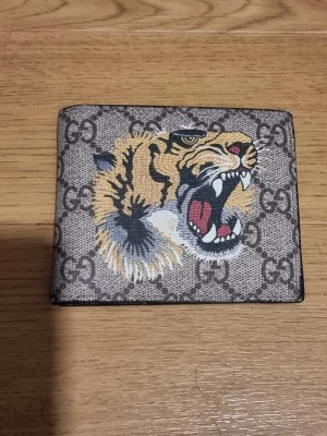 Fin plånbok - INSTABOX!!! Säljer en snygg brun plånbok, som har ett stort, färgstarkt tigertryck på framsidan. Plånboken är i canvasmaterial och har en stilren, fyrkantig form. Perfekt för dig som vill sticka ut med en unik accessoar. LÅDA OCH PÅSE INGÅR INTE.