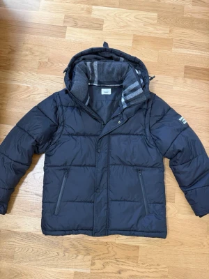 Svart pufferjacka med huva - Snygg svart pufferjacka från burberry med avtagbar huva och armar  dragkedjeförsedda fickor. Insidan har ett mjukt, grått foder med randigt mönster vid kragen. Jackan har en klassisk quiltad design och stängs med både dragkedja och knappar. Perfekt för kalla vinterdagar.