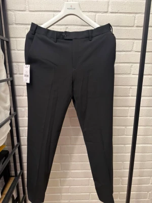 Svarta kostymbyxor 32L - Snygga svarta kostymbyxor med klassisk rak passform och pressveck. Byxorna har hällor för bälte och stängs med knapp och dragkedja. Perfekta för en stilren look och passar till skjorta eller kavaj.