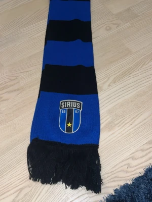 Sirius blå och svart halsduk - Säljer en blå och svart randig halsduk med Sirius klubbmärke och fransar i ändarna. Perfekt för dig som vill visa ditt stöd på matchen eller bara gillar snygga accessoarer med sportig vibe. Stickad i mjukt material och har tydliga klubbfärger.