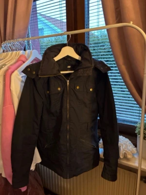 Svart parkasjacka med fuskpäls - Snygg svart jacka från H&M med dragkedja framtill, stora fickor och knappar vid bröstet. Jackan har en hög krage och huva med avtagbar fuskpäls i brunt och beige. Perfekt för kyliga dagar och enkel att matcha med olika outfits. 🌸