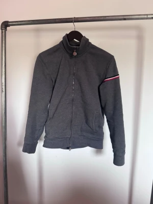 Grå zip-hoodie från Moncler - Snygg grå zip-hoodie från Moncler med hög krage och dragkedja hela vägen. Ärmen har en röd, vit och blå rand samt Moncler-logga. Två fickor framtill och ribbade muddar. Perfekt för en chill och stilren look.