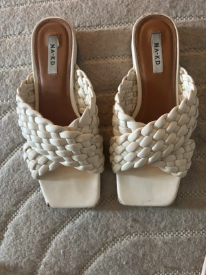 Vita flätade sandaler från NA-KD - Säljer ett par trendiga vita sandaler från NA-KD med breda, flätade remmar och fyrkantig tå. Sandalerna har öppen häl och är tillverkade i ett konstlädermaterial. Perfekta för en stilren och modern look.