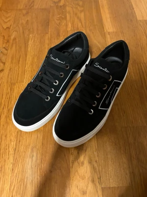 Santoni sneakers HELT NYA - Snygga svarta sneakers från Santoni med vit sula och vita detaljer på sidan. Skorna har svarta skosnören, metallöljetter och en kontrasterande orange detalj under sulan. 